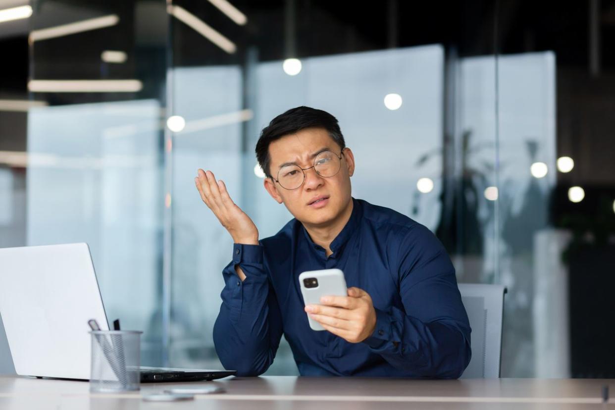 An angry young man in glasses staring confusedly at his phone.