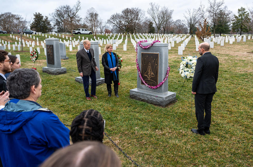 three men in suits stand near a large gray tombstone in a cemetery, with smaller white markers stretching into the distance.