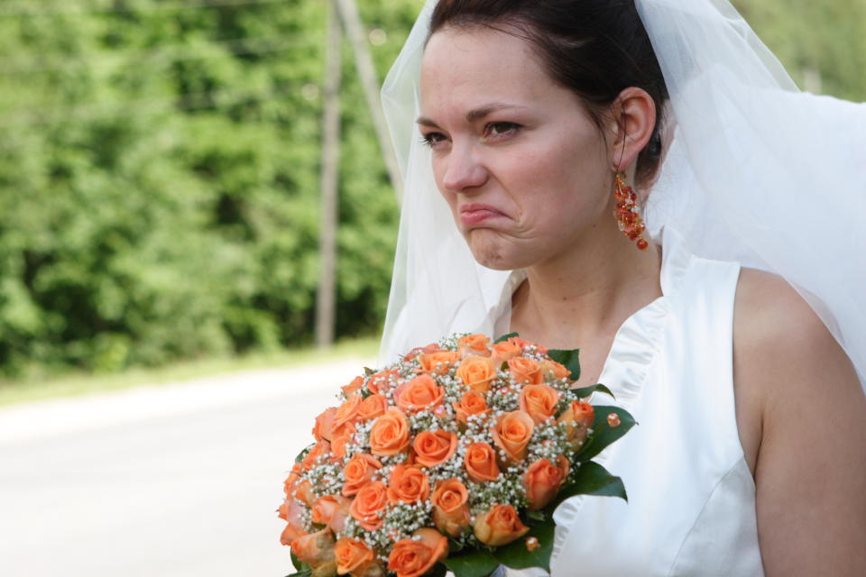 An unhappy bride wearing a white dress and holding a bouquet of orange roses while making an angry expression