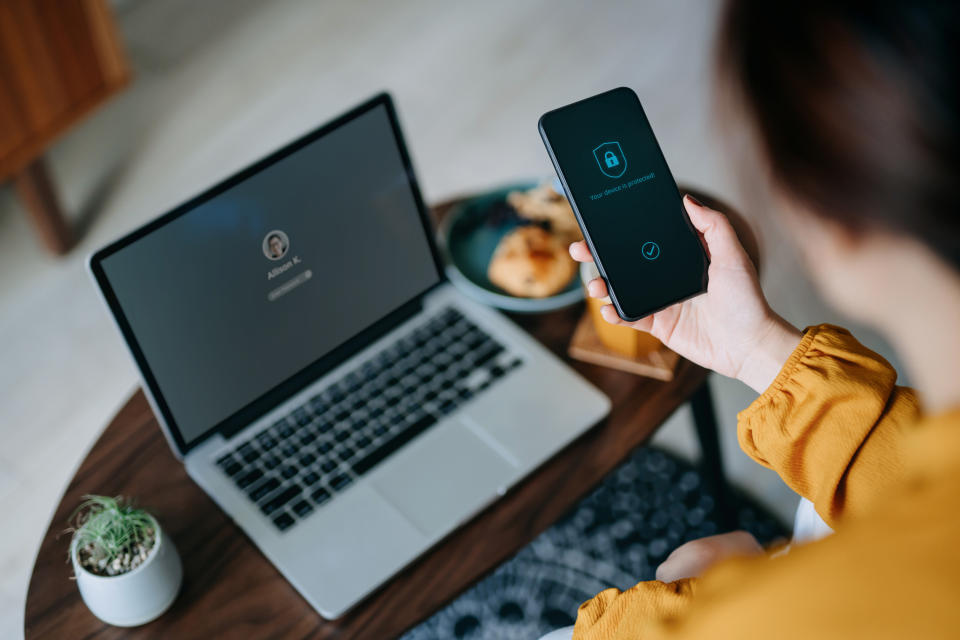 Young Asian woman logging in to her laptop and holding smartphone on hand with a security key lock icon on the screen, sitting in the living room at cozy home. Privacy protection, internet and mobile security concept