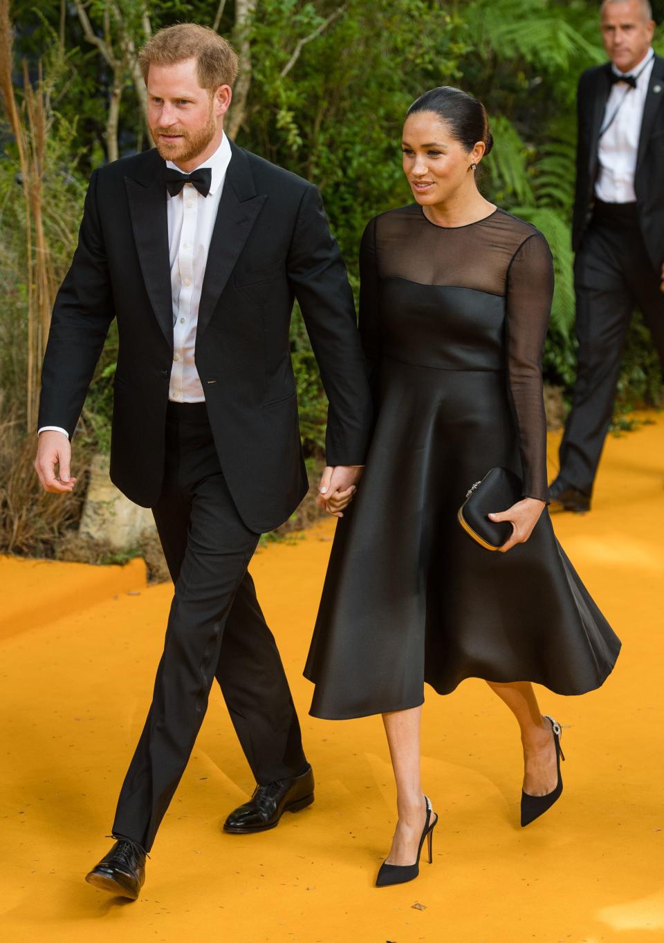 Harry in a black suit and bowtie and Meghan in a black aline dress with a mesh neckline and sleeves.