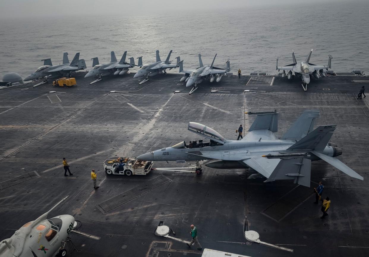 Sailors moving a fighter jet on the flight deck of an aircraft carrier, with other jets lined up on the deck behind it.