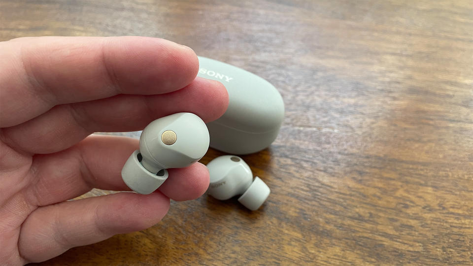 Sony WF-1000XM5 in white with one earbud held in front of the carry case and the other bud on a table