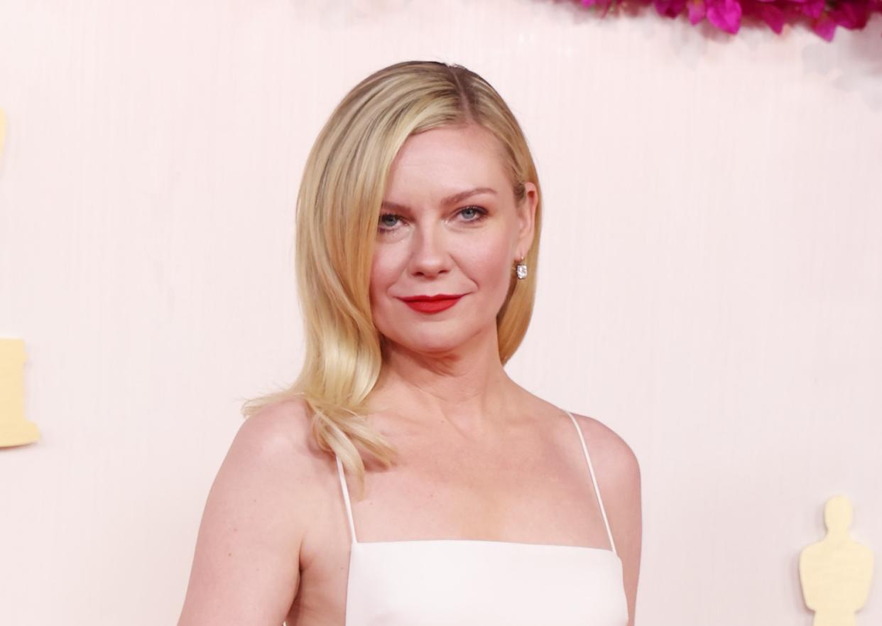  A headshot of Kirsten Dunst on a red carpet wearing a white dress and smiling at the camera. 