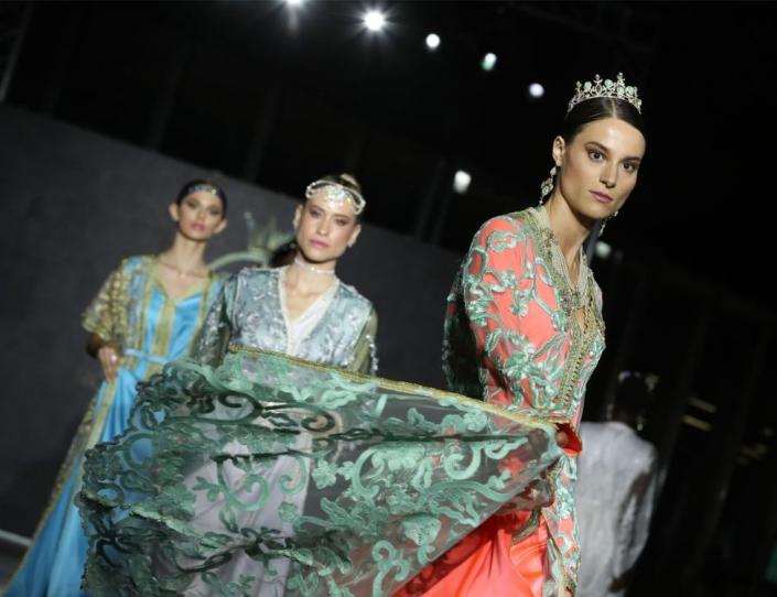 Three models in long-sleeved gowns walk down a runway.