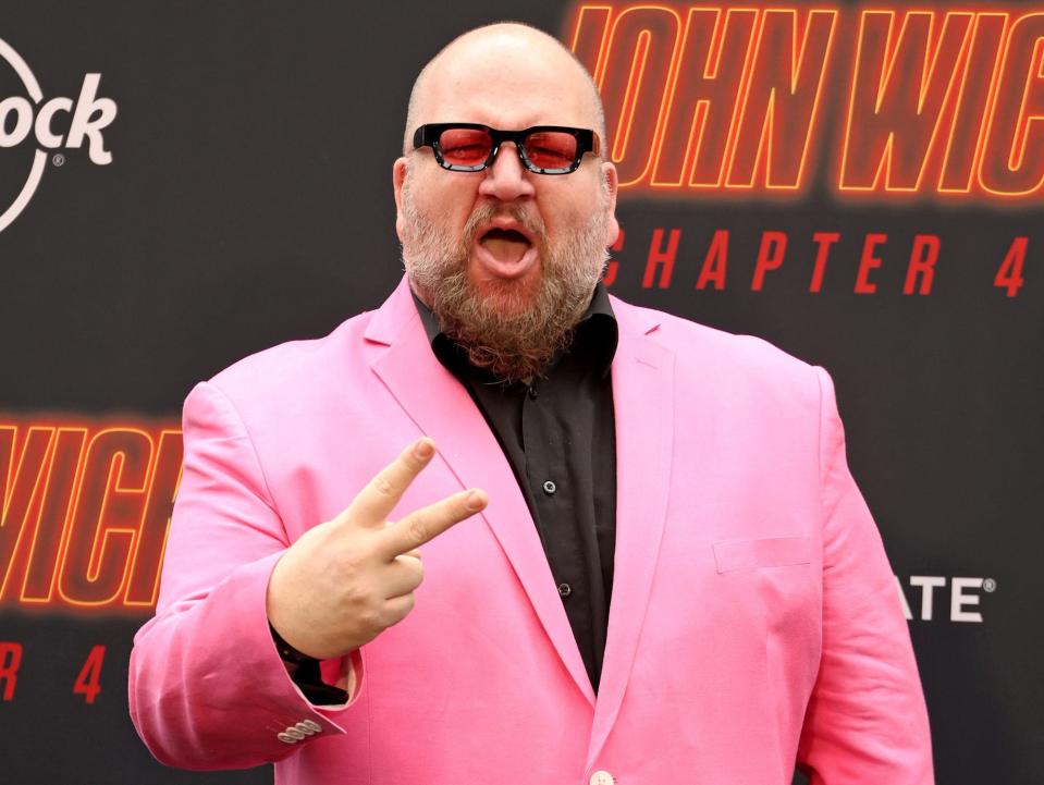 Stephen Glickman wearing a light pink blazer, a black button-down shirt, and a pair of glasses while holding up his fingers and exclaiming