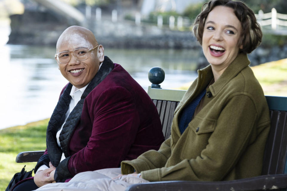 Two actors sitting on a bench, one in a red velvet jacket and tie, the other in a green coat, both smiling