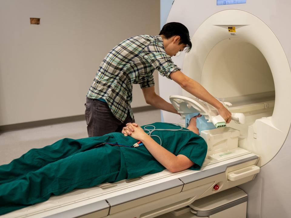 A researcher places a grid on the head of a participant in blue scrubs, who is lying down ready to enter an fMRI machine.