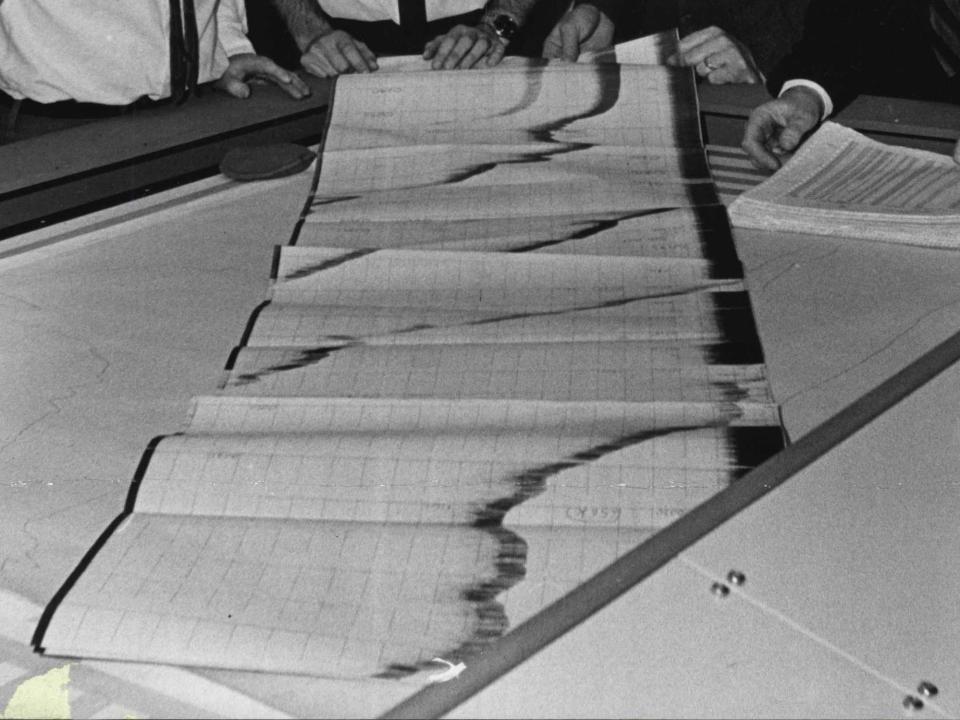 Peope stand around a large echo sounding machine with a large piece of paper and depth data