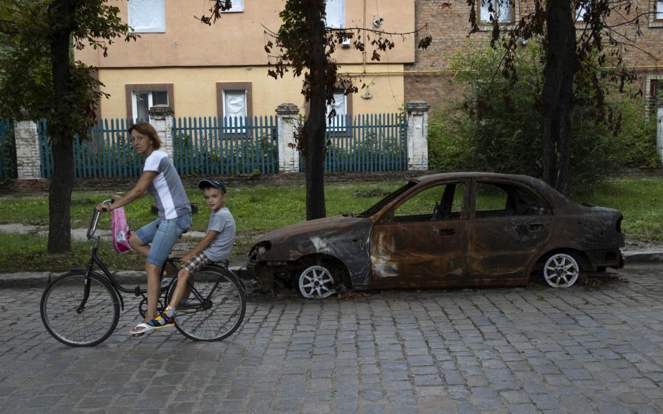 A Ukrainian mother and child ride a bike past a car destroyed by Russian shelling in Orikhiv, Zaporizhzhia Oblast - Metin Aktas/Anadolu Agency