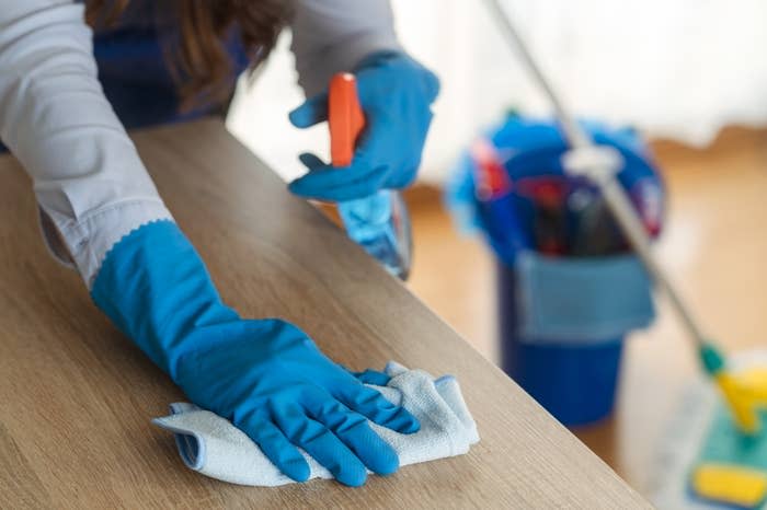 A person wearing blue gloves cleans a wooden table with a cloth, holding an orange spray bottle. A mop and a bucket with cleaning supplies are visible in the background