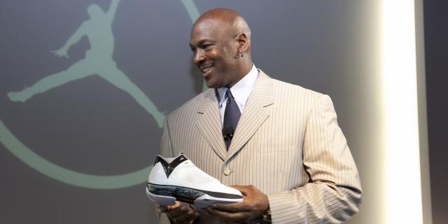 michael jordan, wearing a tan pinstriped suit and black tie, smiles and holds a sneaker while standing in front of the nike air jordan logo