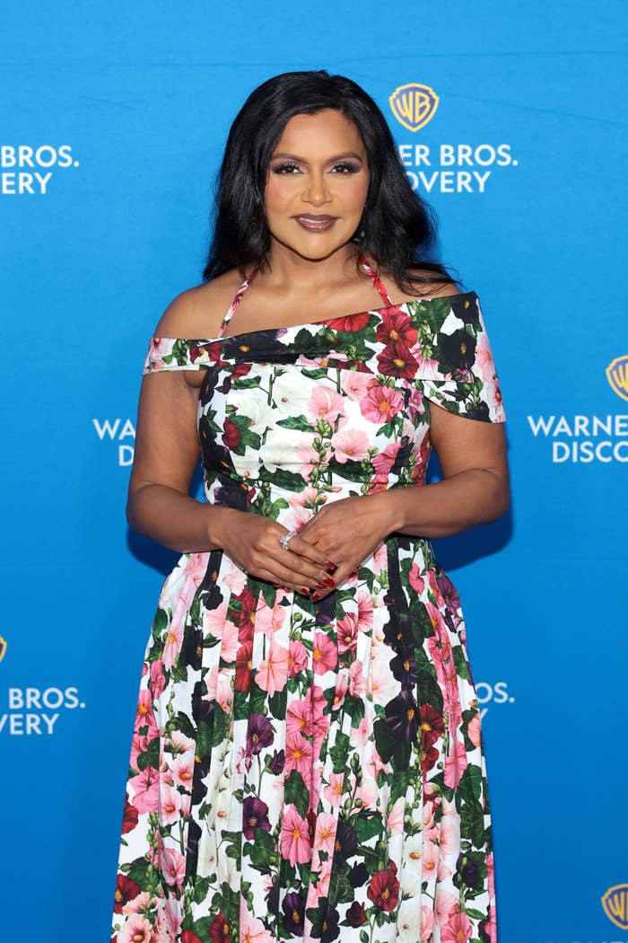 Mindy Kaling stands on the Warner Bros. Discovery red carpet, wearing a floral off-shoulder dress and smiling at the camera