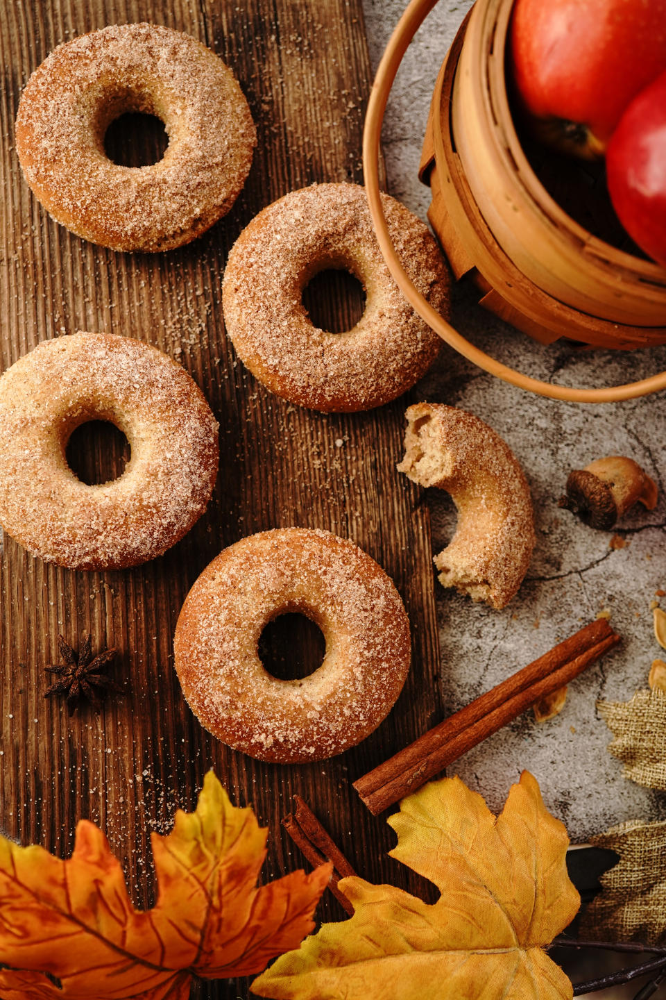 Apple cider donuts sprinkled with sugar are placed on a wooden board next to apples in a basket, a cinnamon stick, and colorful autumn leaves