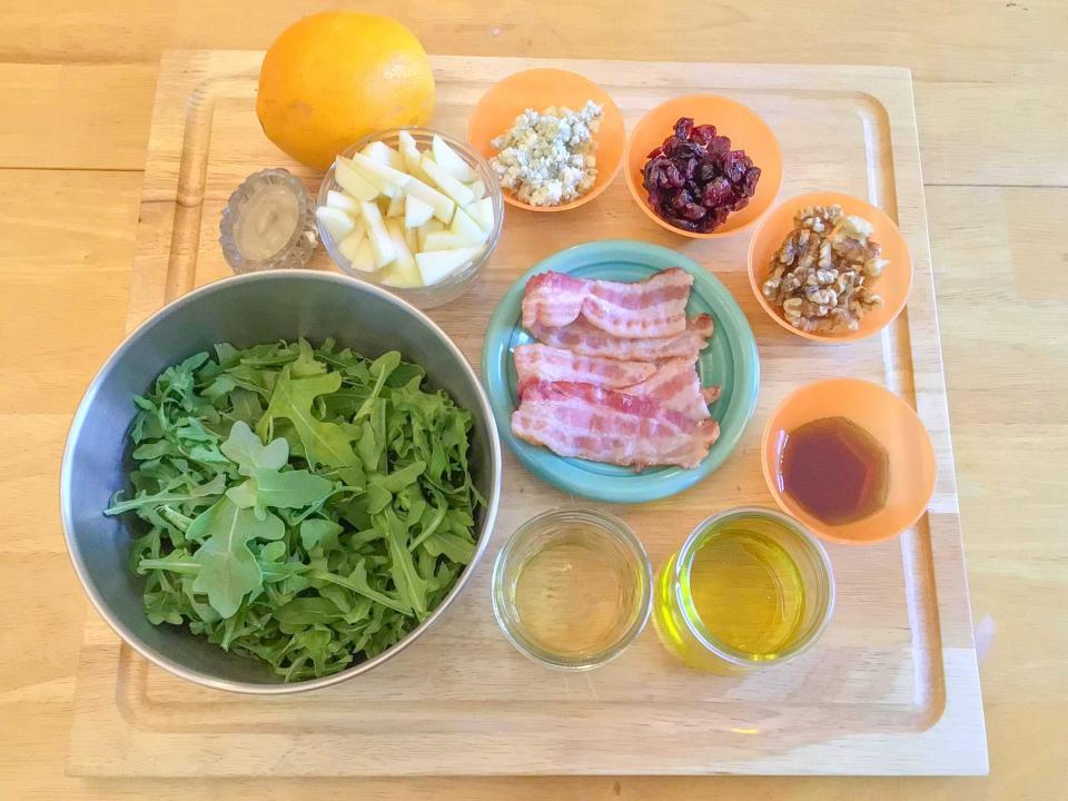 A wooden cutting board holding bowls of ingredients for a Cobb salad. The ingredients include arugula, bacon, oil, cranberries, nuts, an orange, and blue cheese.