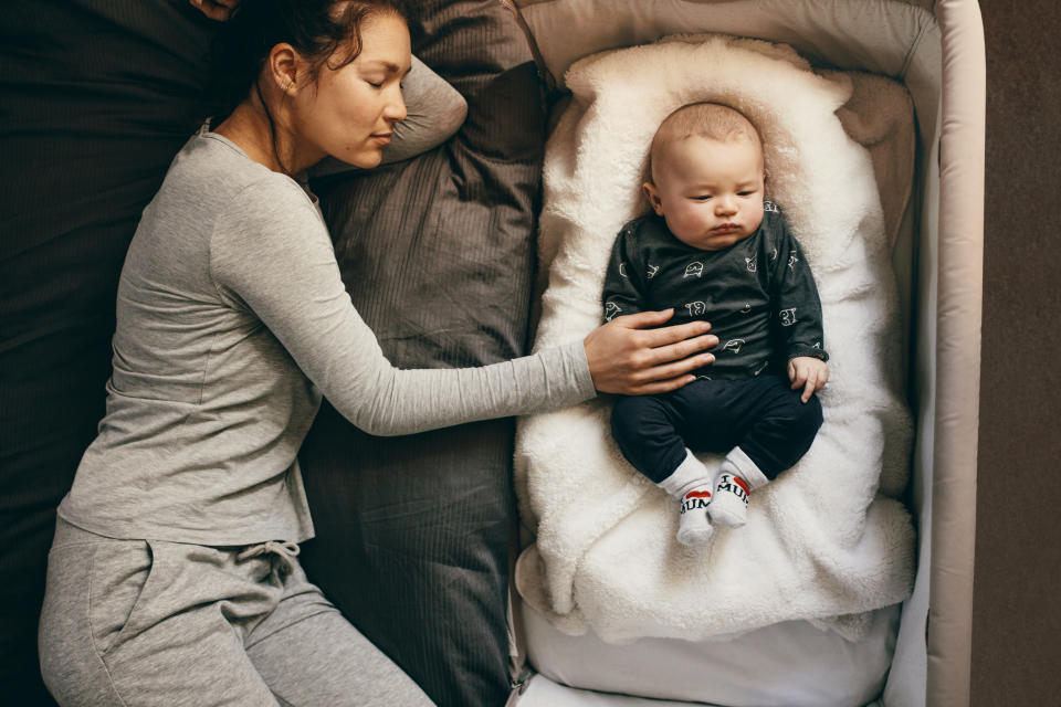 A woman in gray sleepwear rests on a bed beside a baby lying in a bassinet. The woman gently places her hand on the baby's chest as they both sleep