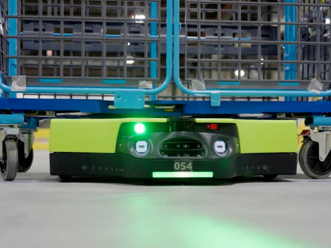 A green wheeled robot carries a large, wheeked cage on its back.