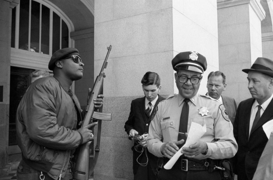 Lt. Ernest Holloway reads a document as a member of the Black Panther Party in a beret stands to attention, his rifle pointed in the air.