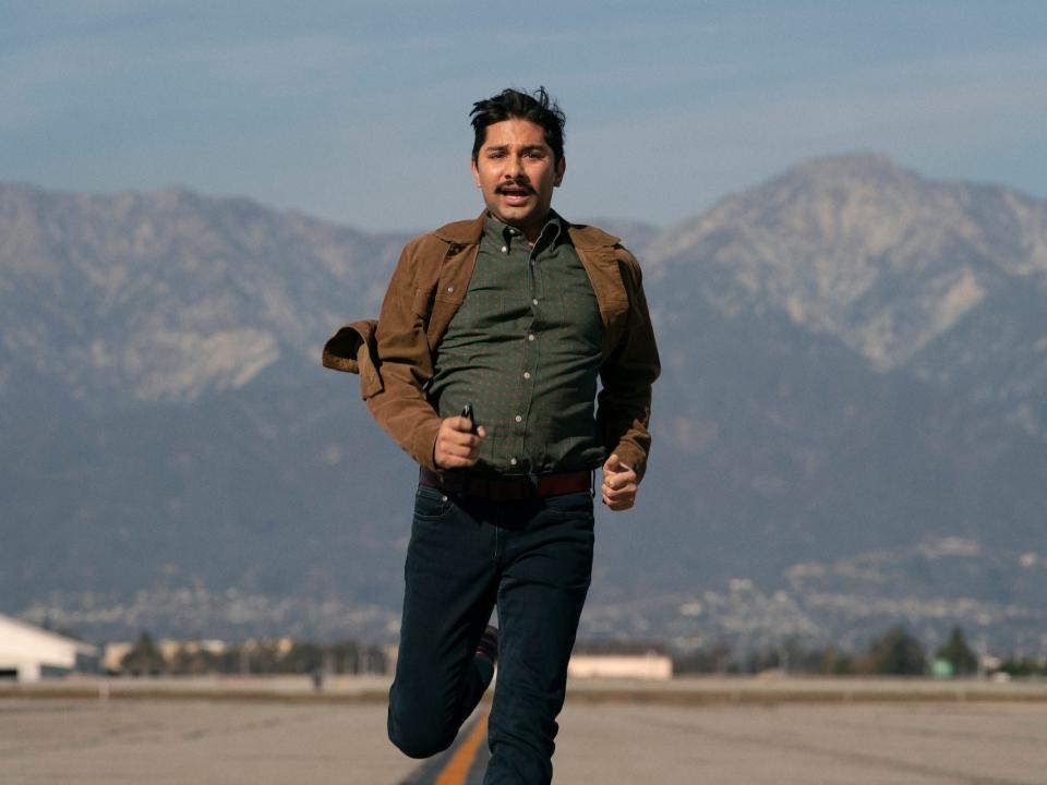 mark indelicato as damien in hacks, wearing a green shirt, brown jacket, and jeans, clutching a pair of sunglasses as he runs down an airplane tarmac