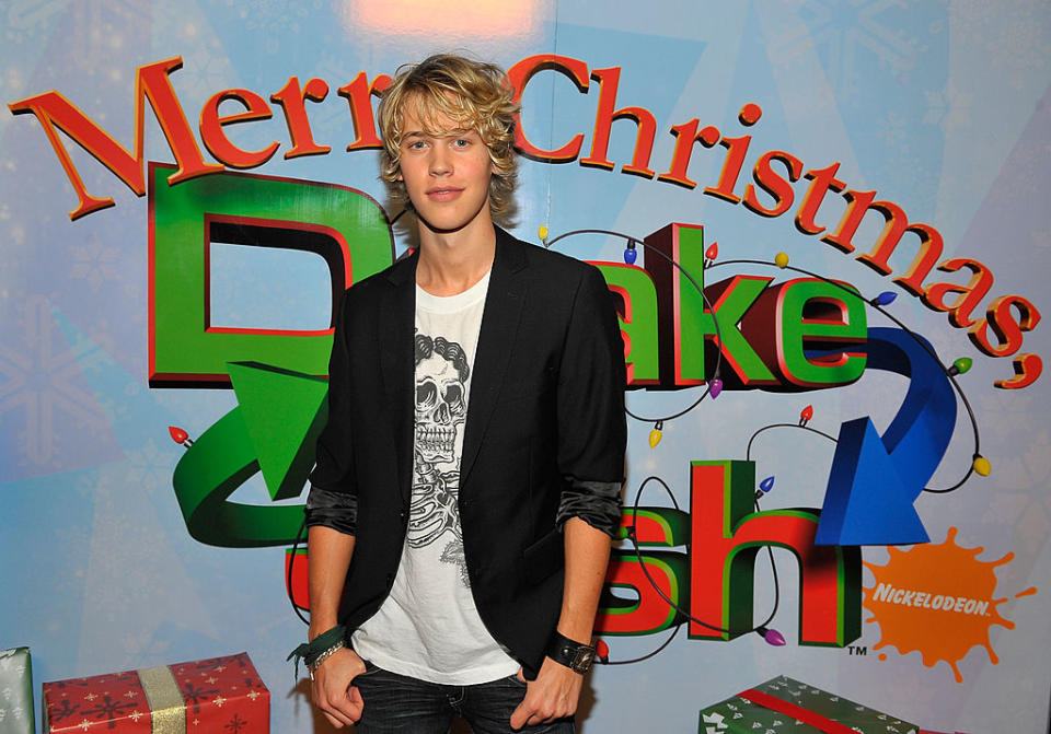 Person standing before a 'Merry Christmas, Drake & Josh' backdrop, wearing casual graphic tee and necklace