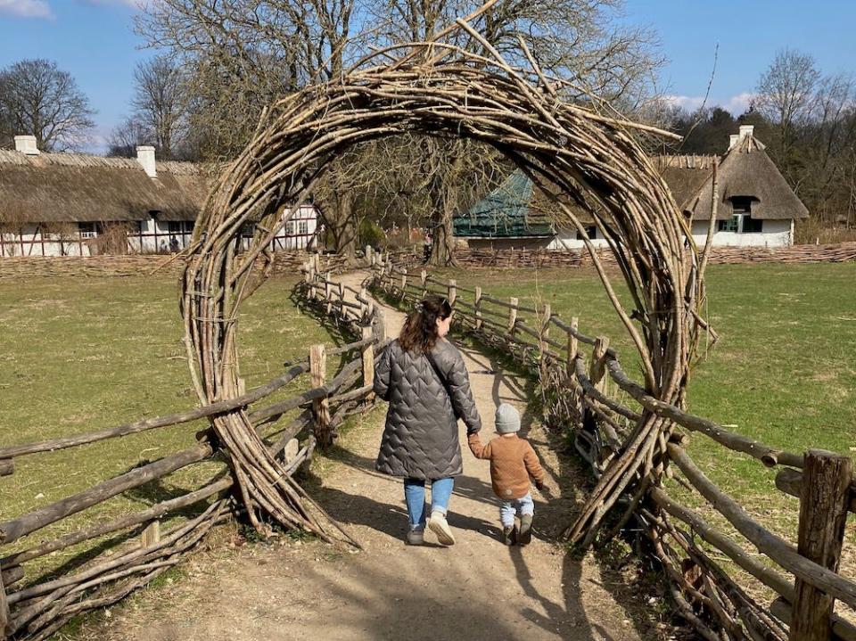 Picture from behind of Ilana Buhl holding hands with her son as they walk down a dirt path outside. Ilana wears a brown winter puffy jacket, blue jeans, and white sneakers. Her son wears a grey knit beanie, tan jacket, blue pants, and brown shoes. Above them is a circular archway made from branches tied together. On either side of them is a crudely made wooden fence. In front of them are several low white houses with thatched roofs. 