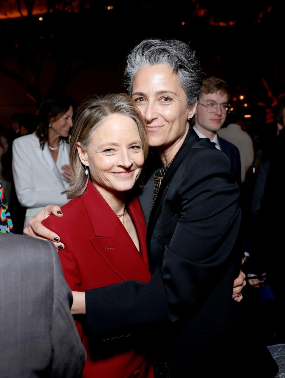 Jodie Foster and Alexandra Hedison embrace at an event, both wearing suits. Jodie wears a red suit, and Alexandra wears a black suit. People are seen in the background