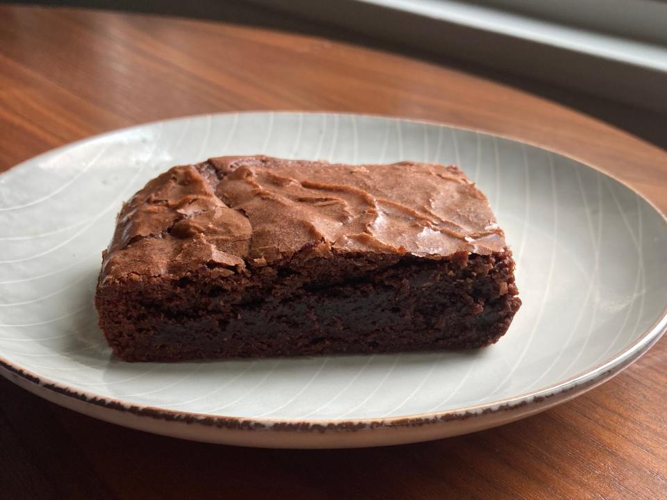 A flatter, light-brown brownie sits on a white plate on a wooden table