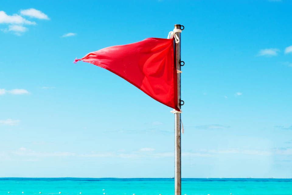 A red flag waves gently on a pole against a backdrop of a clear ocean and blue sky