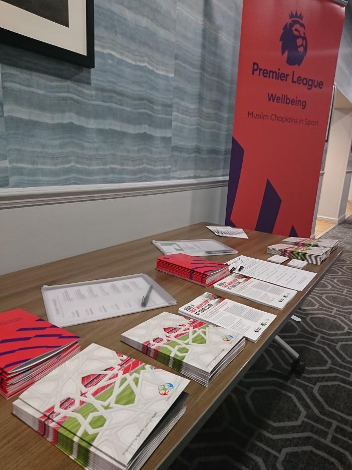 A picture of some leaflets and literature from the Muslim Chaplains In Sport, on a light brown table with an orange Premier League sign next to it that reads 'Premier League Wellbeing - Muslim Chaplains in Sport'