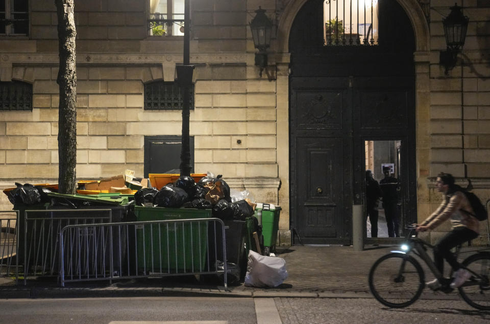Man rides a bicycle past not collected garbage cans at the staff entrance of the Elysee Palace in Paris, Monday, March 13, 2023. A contentious bill that would raise the retirement age in France from 62 to 64 got a push forward with the Senate's adoption of the measure amid strikes, protests and uncollected garbage piling higher by the day. (AP Photo/Michel Euler)