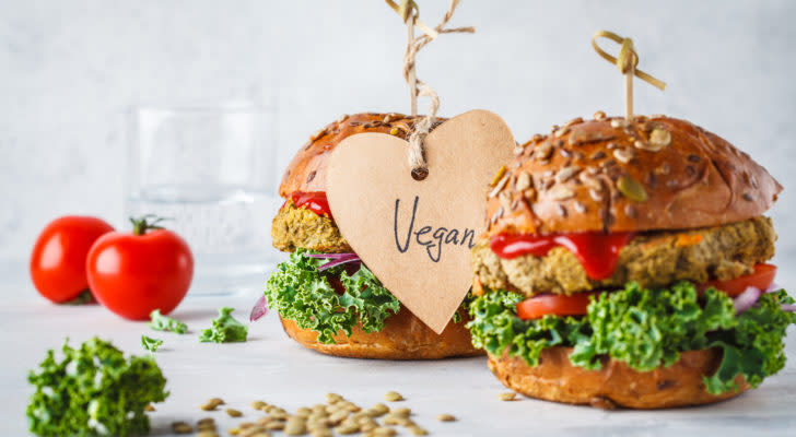 Two plant-based burgers are placed next to a variety of vegetables. One of them has a heart-shaped label hanging off of it with the handwritten word "vegan" on it.