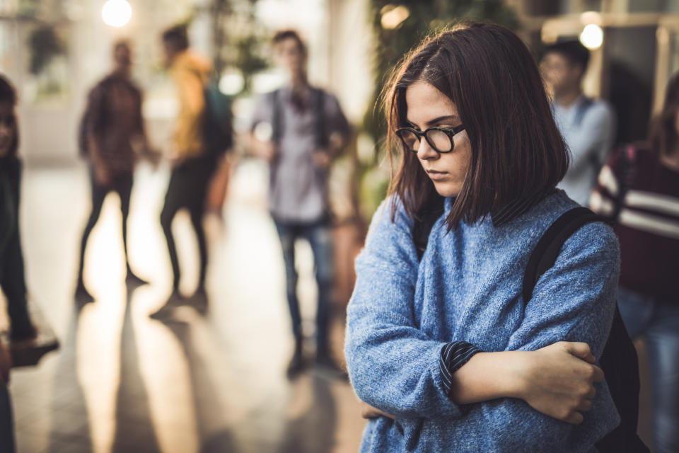 A young girl with glasses and a backpack stands with her arms crossed, looking down. Several blurred figures in the background appear to be talking