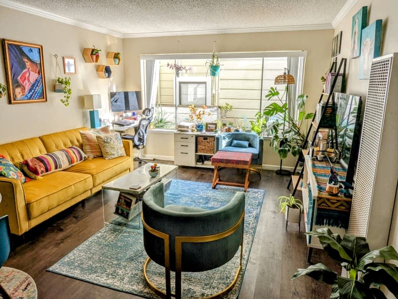 Beige living room with gold sofa, blue curved accent chairs, blue rug, and lots of plants