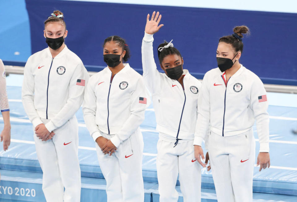 Biles waving as she stands with her teammates on the floor of the arena