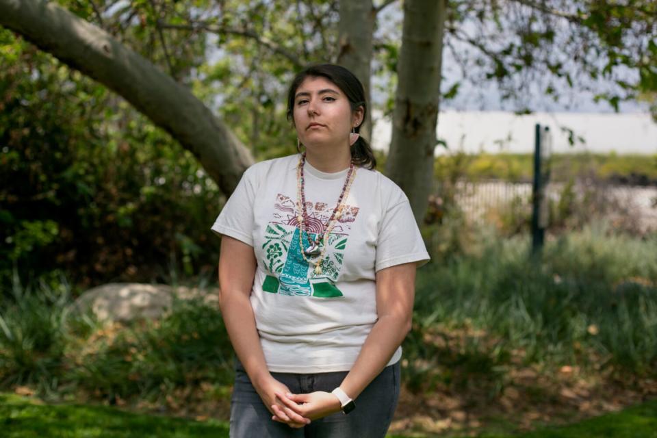 A young woman with dark hair and wearing a T-shirt stands in a shade of a tree behind her.