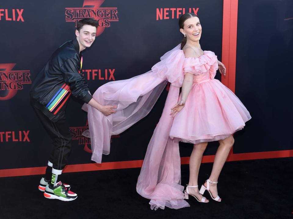 Millie Bobby Brown smiles in a pink dress as Noah Schnapp fluffs her train on Stranger Things red carpet