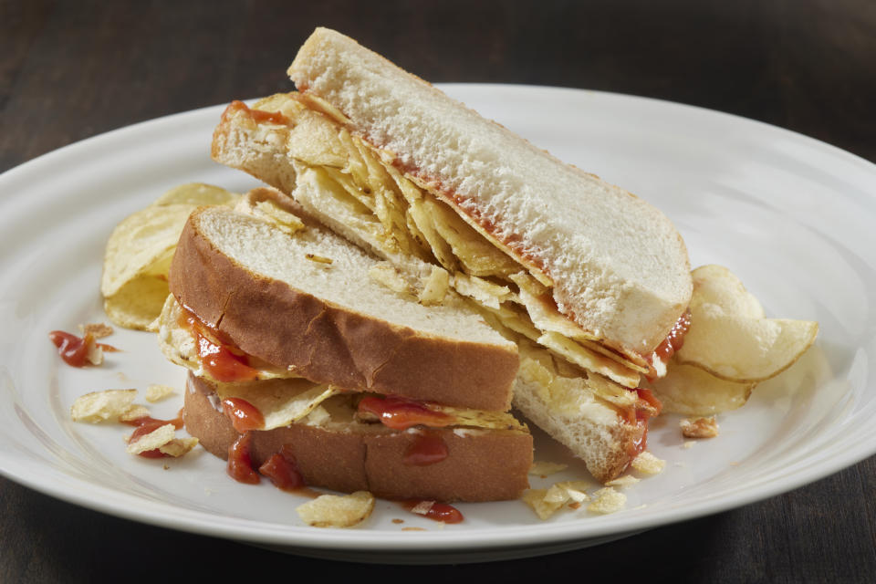 Two halves of a sandwich on white bread with potato chips and ketchup stacked between the slices. Chips scattered around the plate