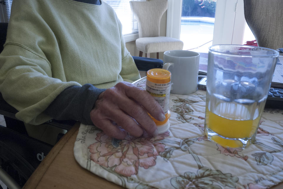 An elderly woman reaches for a pill bottle on a table with a glass of orange juice.