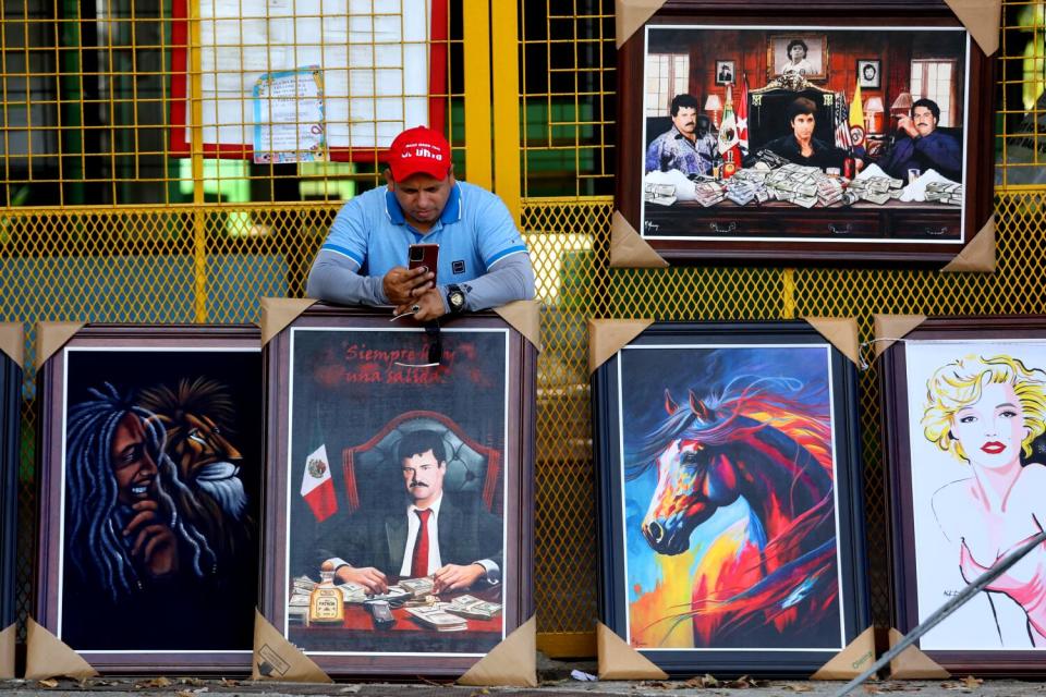 A man in a red cap and blue shirt leans his elbows on a painting of a seated man in a dark suit and red tie, near other works