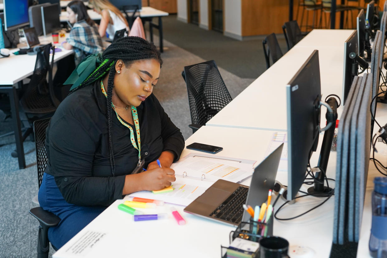 Person wearing sunflower lanyard to show less visible disabilities sits at desk writing on post-it note, with coloured highlighters and a notepad on the desk and an open plan office behind them.