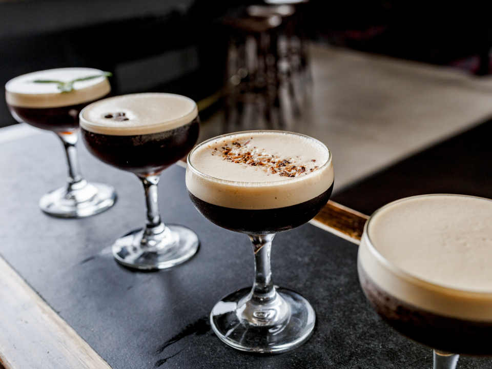 Vier Espresso-Martinis mit verschiedenen Garnierungen auf einer Bar aufgereiht.