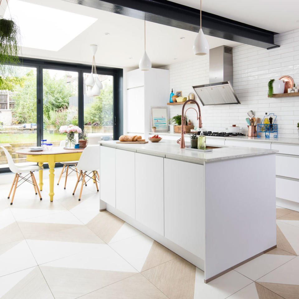 white kitchen with a white island  and a kitchen sink with a brass kitchen tap and a gas stove, next to a wooden yellow table  with white chairs with wooden legs decorated with flowers