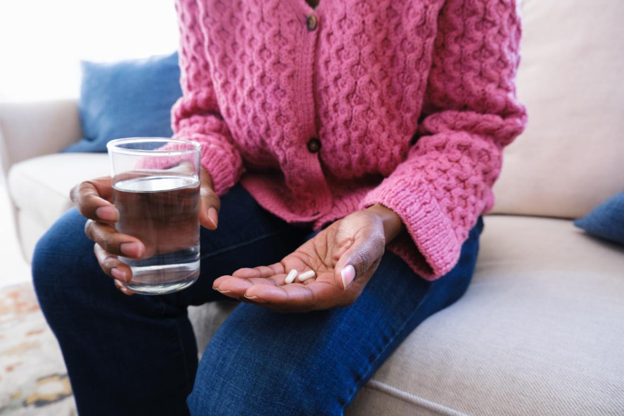 A person wearing a pink cable knit sweater holds a glass of water in one hand and supplements in the other.