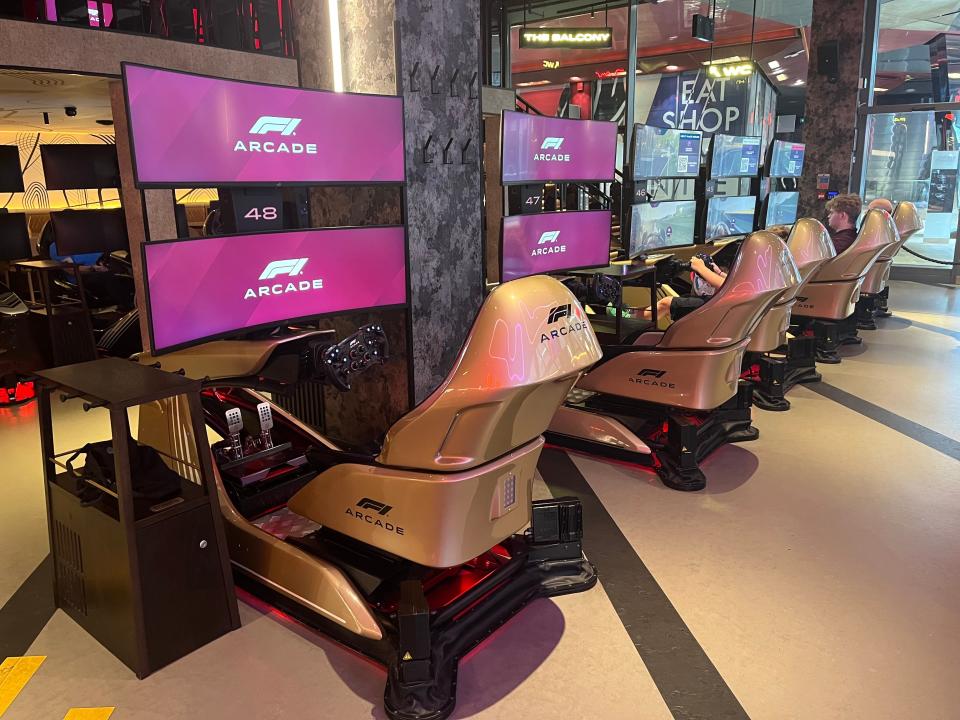 F1 Arcase Driving simulator machines in a row, which have two screens, a steering wheel and gold seats.