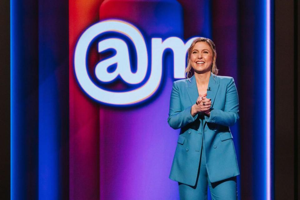 taylor tomlinson smiles while clasping her hands together in front of her chest, she stands in front of a multicolor background with the logo at sign am and wears a bright blue monochrome suit