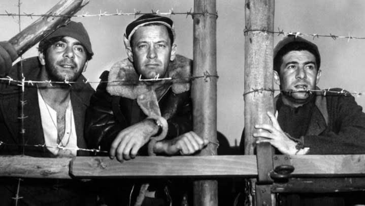 Three men stand behind barbed wire in Stalag 17.