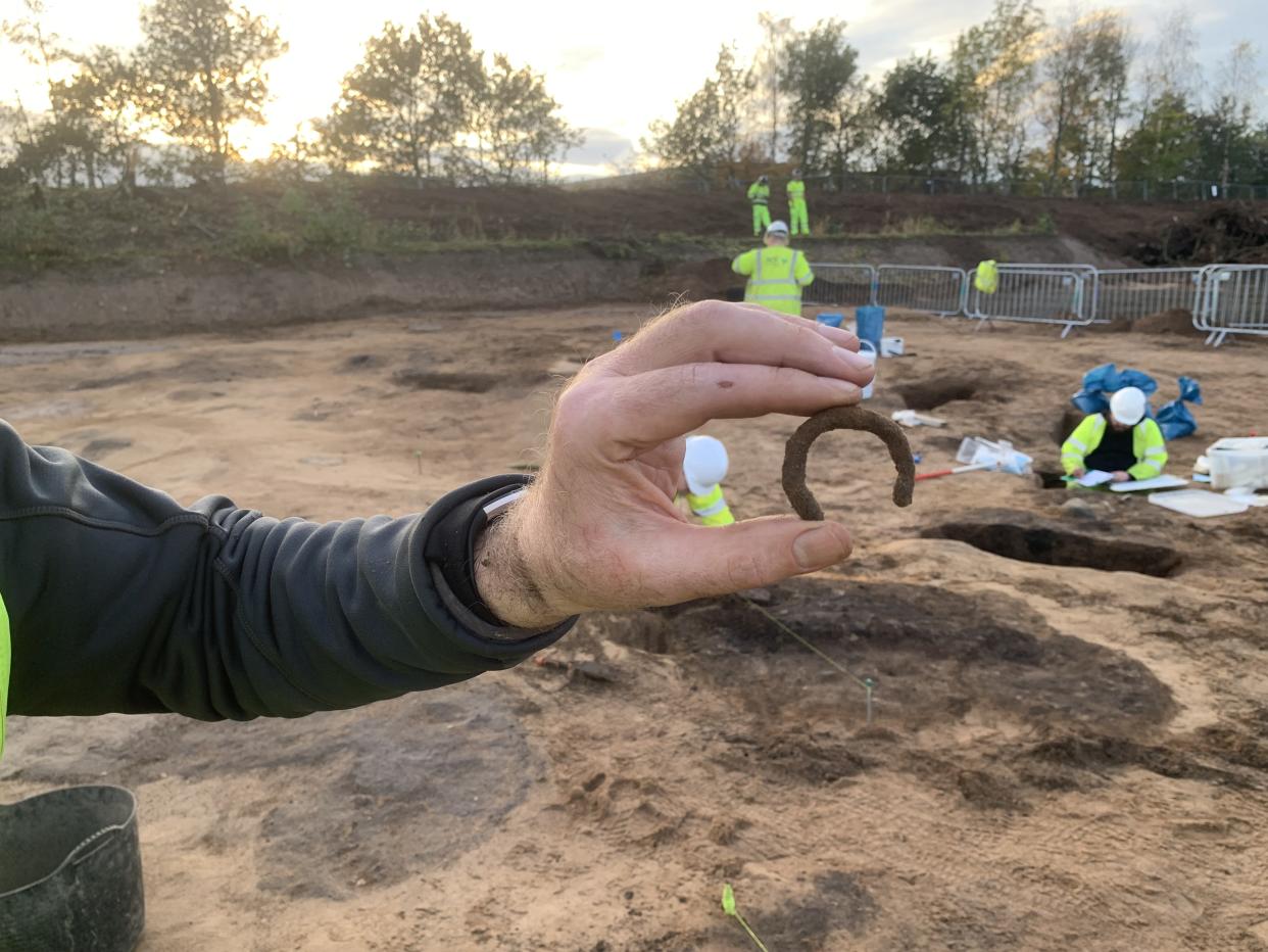 A hand holding a metal ring, with archaeologists in hi-vis clothing in the background in a muddy excavation