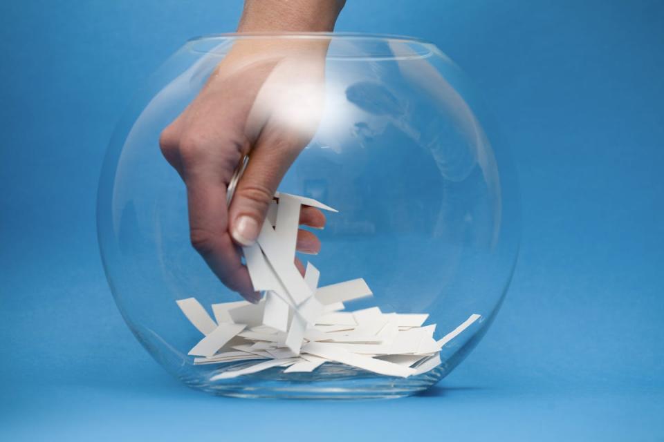 A hand reaches into a glass bowl to choose a piece of paper from a group of them.