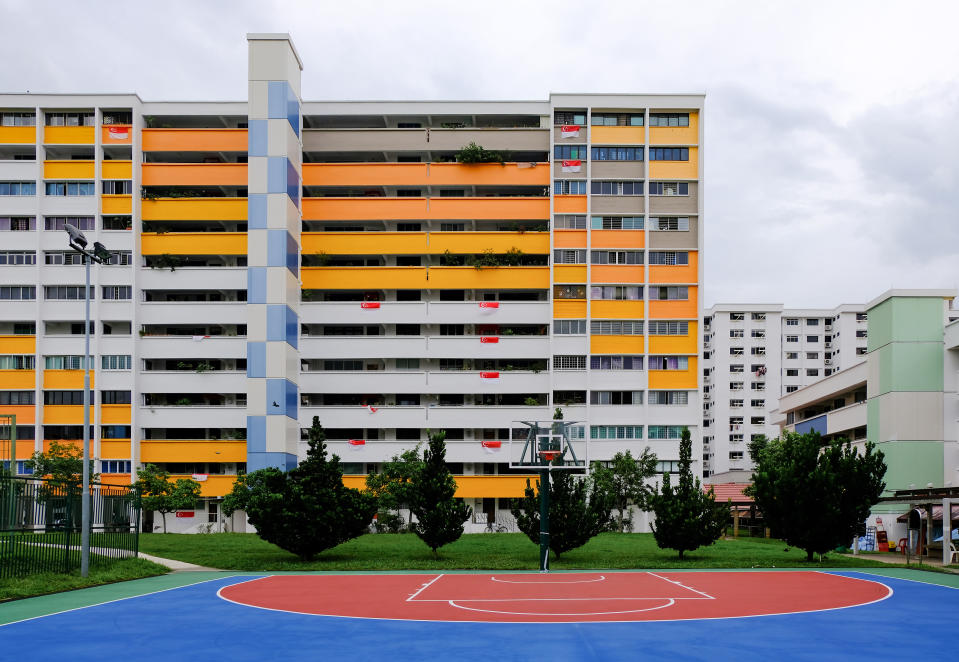Colourful block of HDB flats (painted in white yellow orange blue) in yishun on bright, clear day, Singapore flags hung over some balconies. Neighbourhood park and basketball court in front.