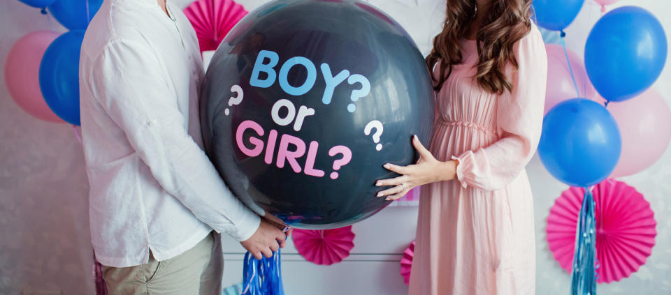 A man in a white shirt and a woman in a pink dress hold a large balloon that reads "BOY? OR GIRL?" amidst blue and pink decorations